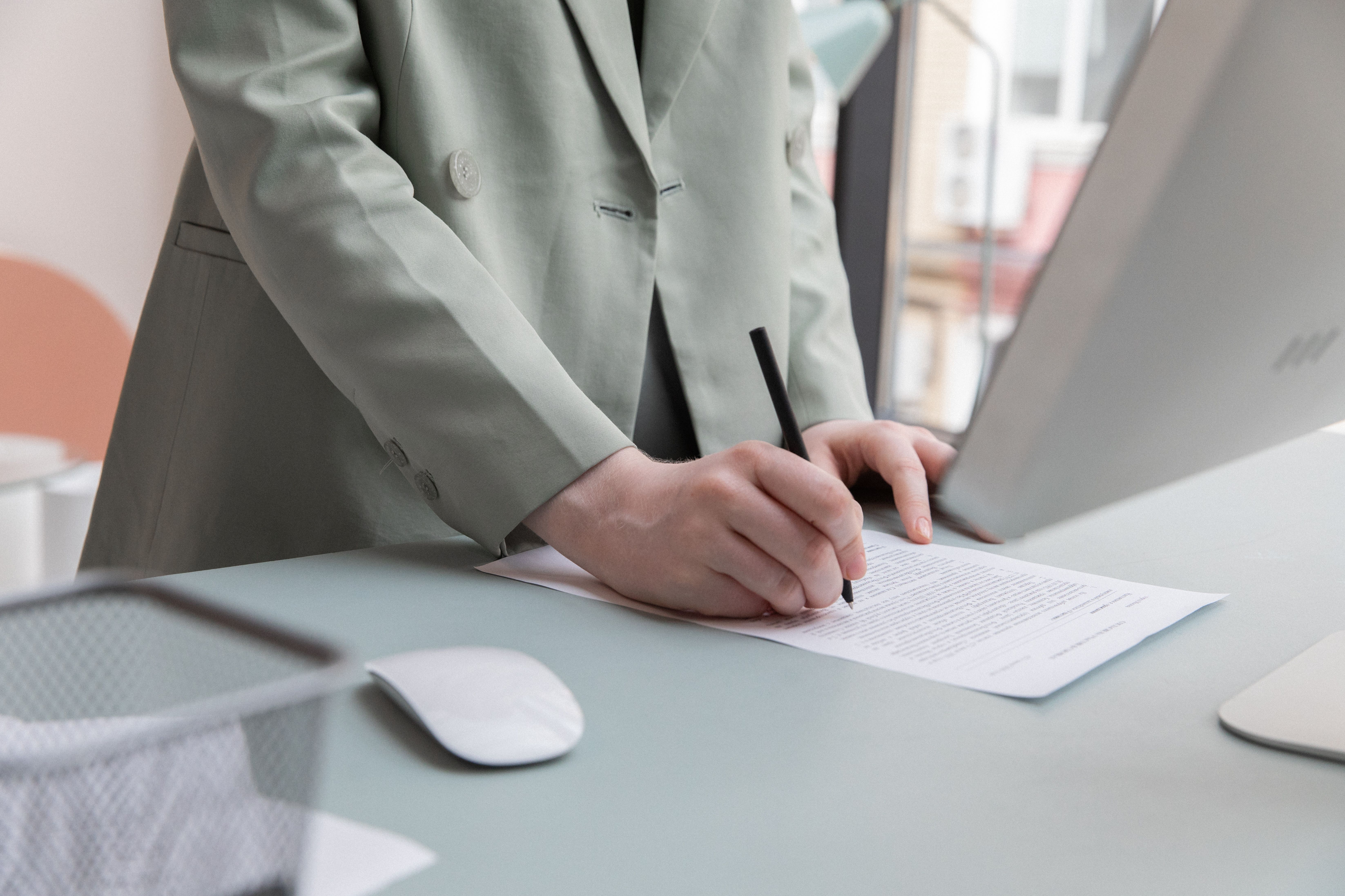 A person in a suit jacket writing on a document at a desk with a computer